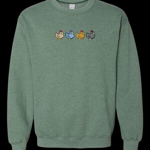 K&ouml;nnte beinhalten: Ein gr&uuml;ner Pullover mit einer Reihe von vier pixelartigen H&uuml;hnern, die auf der Vorderseite gestickt sind. Die H&uuml;hner sind in verschiedenen Farben: rot, blau, gelb und grau.
