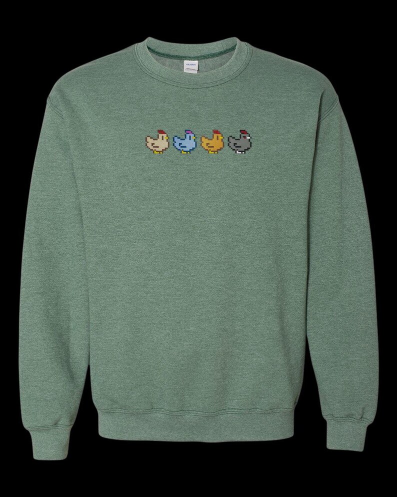 K&ouml;nnte beinhalten: Ein gr&uuml;ner Pullover mit einer Reihe von vier pixelartigen H&uuml;hnern, die auf der Vorderseite gestickt sind. Die H&uuml;hner sind in verschiedenen Farben: rot, blau, gelb und grau.