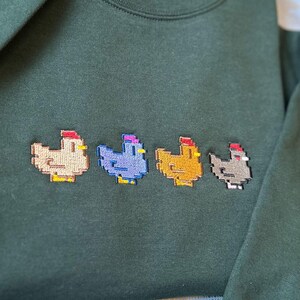 K&ouml;nnte beinhalten: Ein dunkelgr&uuml;ner Pullover mit vier gestickten, pixelartigen H&uuml;hnern in verschiedenen Farben: beige, lila, gelb und grau.