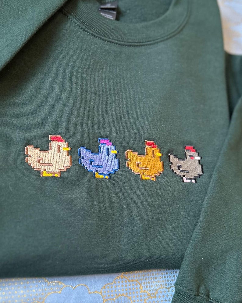 K&ouml;nnte beinhalten: Ein dunkelgr&uuml;ner Pullover mit vier gestickten, pixelartigen H&uuml;hnern in verschiedenen Farben: beige, lila, gelb und grau.