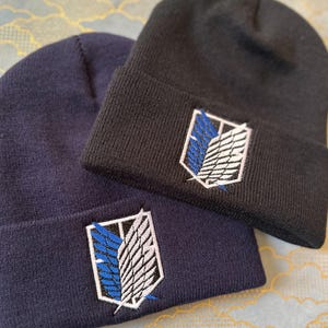 May include: Two knit beanies with the Attack on Titan logo embroidered on them. One beanie is navy blue and the other is black.