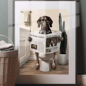 May include: Framed print showing a German Shorthaired Pointer wearing spectacles and reading a newspaper on a toilet. The newspaper's headline reads "Reading Matters". A cactus in a white pot is to the right of the dog.
