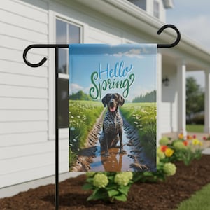 May include: A garden flag with a German Shorthaired Pointer dog on a muddy path. The flag reads "Hello Spring" in script, set against a field and blue sky. The flag is displayed outside a house.
