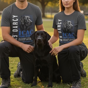 May include: Two charcoal gray t-shirts with the text "K9 Darcy" and "ICAC Task Force" printed in white and blue. A black Labrador Retriever sits between the people wearing the shirts. The shirts have a crew neck and short sleeves.