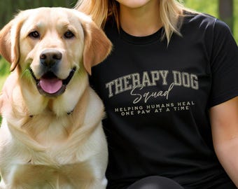 Könnte beinhalten: Eine Frau trägt ein schwarzes T-Shirt mit dem Text "Therapy Dog Squad" und den Worten "Helping Humans Heal One Paw At A Time" in goldenen Buchstaben. Sie steht neben einem Golden Retriever.