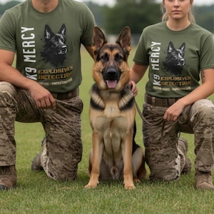 May include: A German Shepherd dog sits between two people wearing olive green t-shirts with the text "K9 Mercy" and a graphic of a German Shepherd. The shirts also read "Explosives Detection". The people are kneeling on the grass.
