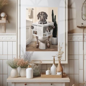 May include: A framed print shows a dog wearing spectacles, reading a newspaper on a toilet. The dog is brown and white. The newspaper headline reads "Morning Routine". The bathroom setting includes a cactus and a towel rail. The print is in a white frame.