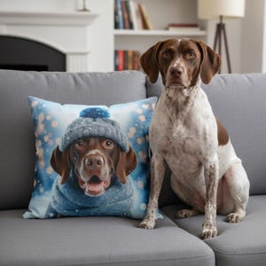 May include: A decorative square pillow featuring a dog wearing a blue knit hat and scarf, set against a snowy background. A real dog with brown and white fur sits next to the pillow on a gray couch.