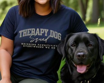 Könnte beinhalten: Eine Person trägt ein marineblaues T-Shirt mit dem Text "Therapy Dog Squad" und den Worten "Bringing Pawsitivity Everywhere We Go". Ein schwarzer Labrador Retriever sitzt neben der Person.