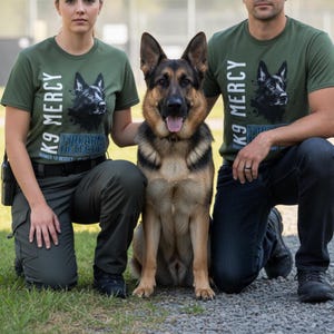 May include: A German Shepherd dog sits between two people wearing olive green t-shirts. The shirts feature a black and white dog portrait and the text "K9 MERCY" and "FIREARMS DETECTION". The dog has a black and tan coat.