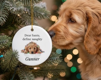 May include: A white, round Christmas ornament with a gold string, featuring a puppy image and the text "Dear Santa, define naughty" and the name "Gunner". The ornament is hanging on a Christmas tree with a golden retriever puppy.
