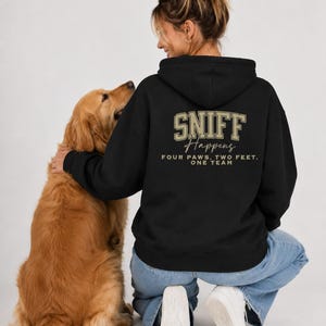 May include: A black hooded sweatshirt featuring gold and white text on the back that reads SNIFF Happens FOUR PAWS, TWO FEET. ONE TEAM. The hoodie has a drawstring hood, long sleeves, and a ribbed hem, worn with light blue denim jeans and white sneakers.
