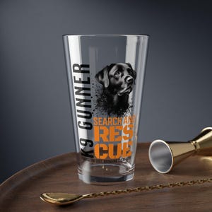 May include: A clear pint glass with a black Labrador Retriever illustration and the words "K9 Gunner Search and Rescue" in black and orange. The glass also includes the phrase "Finding the lost, bringing them home."