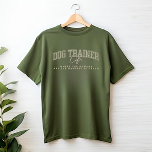 Pode incluir: Camiseta verde-oliva com o texto "DOG TRAINER Life" em uma fonte universitária. Abaixo, o texto diz "WHERE THE HUMANS ARE THE HARDEST TO TEACH". A camiseta está pendurada em um cabide de madeira sobre um fundo branco.