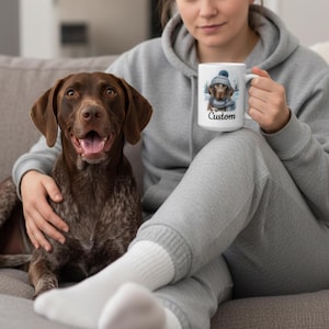 May include: A brown and white German Shorthaired Pointer dog sits next to a person holding a white mug. The mug features a watercolor illustration of a dog wearing a hat and scarf, with the word "Custom" below the image.