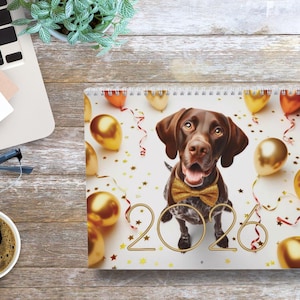 May include: A 2026 calendar featuring a brown dog wearing a gold bow tie, surrounded by gold and orange balloons and confetti. The calendar is on a wooden desk with a laptop, coffee, and glasses.