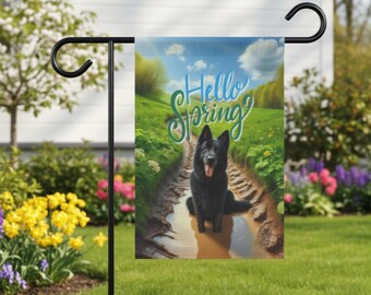 Puede incluir: Bandera de jardín con un perro negro en un camino embarrado y la frase "Hello Spring" en cursiva. El fondo muestra un paisaje verde bajo un cielo azul. La bandera está en un soporte de metal negro.
