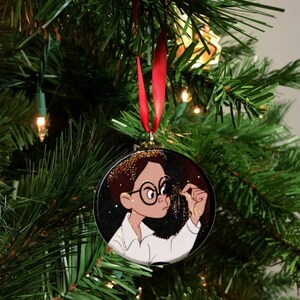 May include: Round Christmas ornament featuring a cartoon illustration of a person with glasses and a white shirt, holding gold glitter. The ornament hangs from a red ribbon against a backdrop of a Christmas tree.