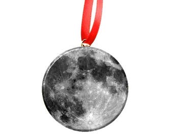 May include: A round, silver ornament with a black and white image of the moon. The ornament is attached to a red ribbon.
