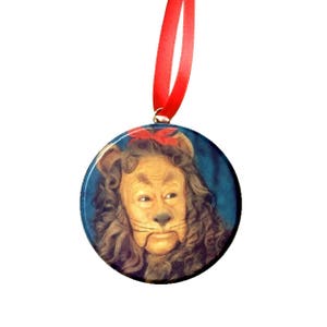 May include: A round, ceramic ornament featuring a portrait of the Cowardly Lion from the Wizard of Oz. The lion is wearing a red bow and has a serious expression on its face.