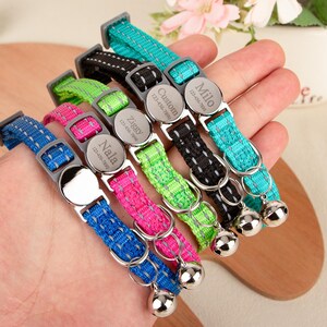 May include: A hand displays six personalized pet collars in various colors: blue, pink, green, black, and teal. Each collar has a silver name tag with a pet's name and phone number, plus a silver bell.