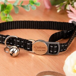 May include: Black cat collar with reflective stitching and a silver bell. The collar features a wooden-style buckle engraved with "Custom" and a phone number. The collar is designed for pets.