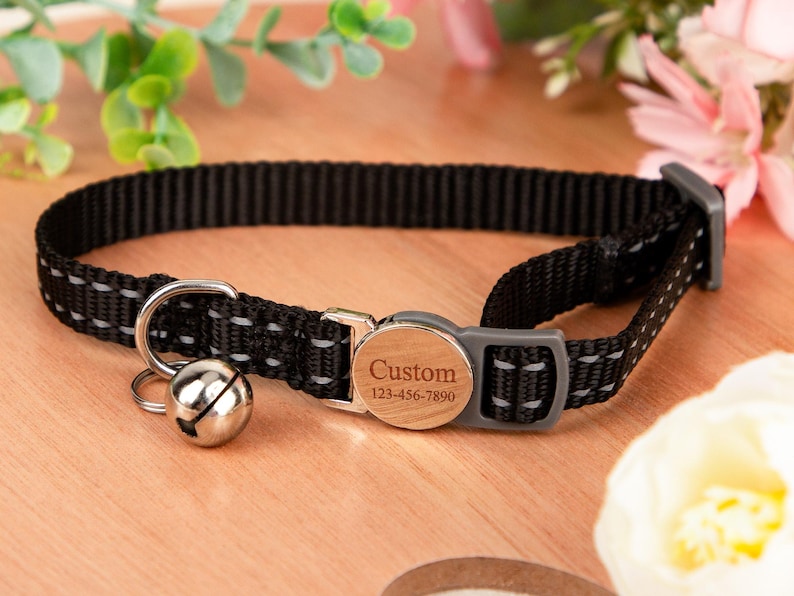 May include: Black cat collar with reflective stitching and a silver bell. The collar features a wooden-style buckle engraved with "Custom" and a phone number. The collar is designed for pets.