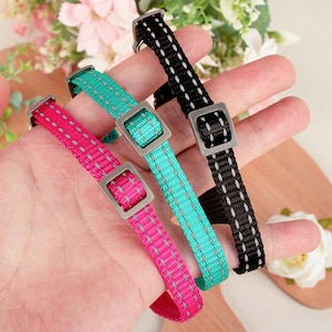 May include: Three colorful dog collars are displayed, each with a square metal buckle. The collars are pink, teal, and black, with reflective stitching. The collars are designed for pets.