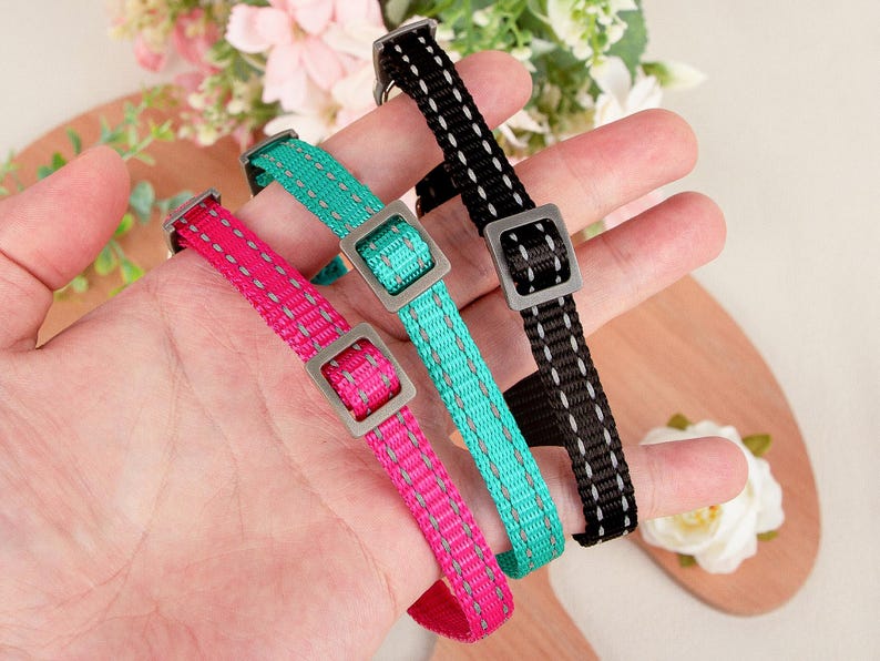 May include: Three colorful dog collars are displayed, each with a square metal buckle. The collars are pink, teal, and black, with reflective stitching. The collars are designed for pets.