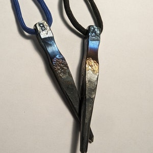 May include: Two metal pendants on cords, one blue and one black. The pendants are shaped like a pointed blade and have a hammered finish. The blue pendant has the letter "B" engraved on it, and the black pendant has the letter "A" engraved on it.