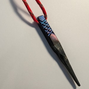 Puede incluir: Una herramienta de metal negro con un cordón rojo y gris adjunto. La herramienta tiene un diseño texturizado y una punta afilada.