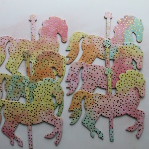 May include: Six pastel-colored paper carousel horses with gold glitter accents. The horses are cut out and ready to be used for crafting projects.