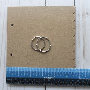 Puede incluir: Un cuaderno de papel kraft con dos anillas plateadas. El cuaderno tiene un borde festoneado y dos agujeros para las anillas. Una regla azul con marcas de cent&iacute;metros se coloca debajo.