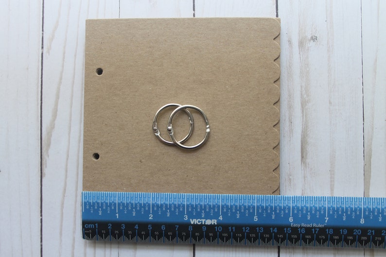 Puede incluir: Un cuaderno de papel kraft con dos anillas plateadas. El cuaderno tiene un borde festoneado y dos agujeros para las anillas. Una regla azul con marcas de cent&iacute;metros se coloca debajo.