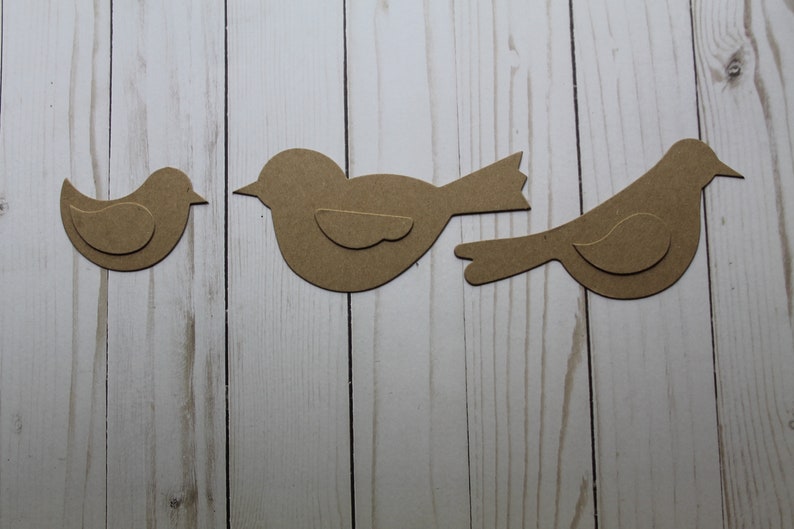 May include: Three brown paper bird cutouts on a white wood background. The birds are different sizes and shapes.