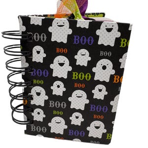 May include: A spiral-bound notebook with a black cover featuring a repeating pattern of white ghosts and the word BOO in various colors. The top of the notebook is decorated with a cluster of purple, orange, and green ribbons and mesh fabric.