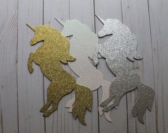 May include: Three glitter unicorn silhouettes in gold, silver, and iridescent white. The unicorns are standing on their hind legs with their horns and tails raised.
