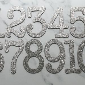 May include: Ten silver glitter numbers, 1 through 10, on a white marble background. The numbers are in a cursive font and are slightly raised.