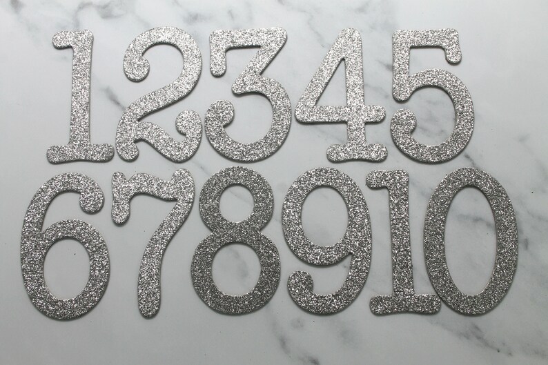 May include: Ten silver glitter numbers, 1 through 10, on a white marble background. The numbers are in a cursive font and are slightly raised.
