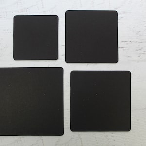 May include: Four black square coasters on a white wooden surface.