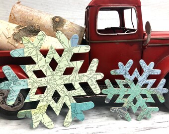 May include: Two paper snowflake ornaments with a map design. The larger snowflake is green and blue, and the smaller snowflake is yellow and blue. The snowflakes are in front of a red toy truck.