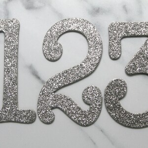 May include: Silver glitter numbers 1, 2, and 3 on a white marble background.