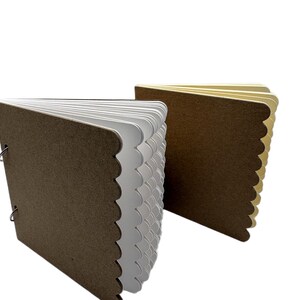 Puede incluir: Dos cuadernos peque&ntilde;os hechos a mano. Las cubiertas son marrones con un borde festoneado. Las p&aacute;ginas son blancas y beige, y los libros se sujetan con anillas met&aacute;licas. Sobre un fondo blanco.
