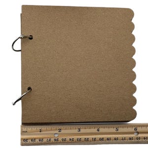 Puede incluir: Un peque&ntilde;o &aacute;lbum de recortes marr&oacute;n con bordes festoneados y anillas met&aacute;licas. Una regla de madera con marcas en pulgadas se coloca junto al &aacute;lbum. El &aacute;lbum mide aproximadamente 20 cm de largo.
