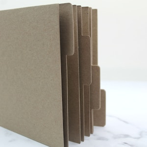 May include: A brown cardboard scrapbook with metal rings and dividers. The scrapbook is closed and standing upright on a white surface.
