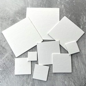 May include: A set of nine white square paper cards of various sizes. The cards are arranged in a random pattern on a gray textured surface.