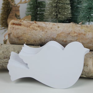 May include: A stack of white paper dove shapes, each with a slightly different design, are arranged on top of two birch logs. The logs are stacked on a white surface.