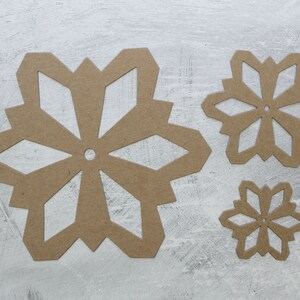 May include: Three brown paper snowflake cutouts of varying sizes are arranged on a textured, light gray surface. The snowflakes have intricate geometric designs with six points and a central hole, perfect for winter crafts and decorations.