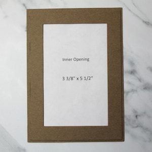 May include: A rectangular brown paper mat with a white inner opening. The text "Inner Opening" is above the dimensions "3 3/8" x 5 1/2"" in the center of the white space. The mat is on a white and gray marble surface.