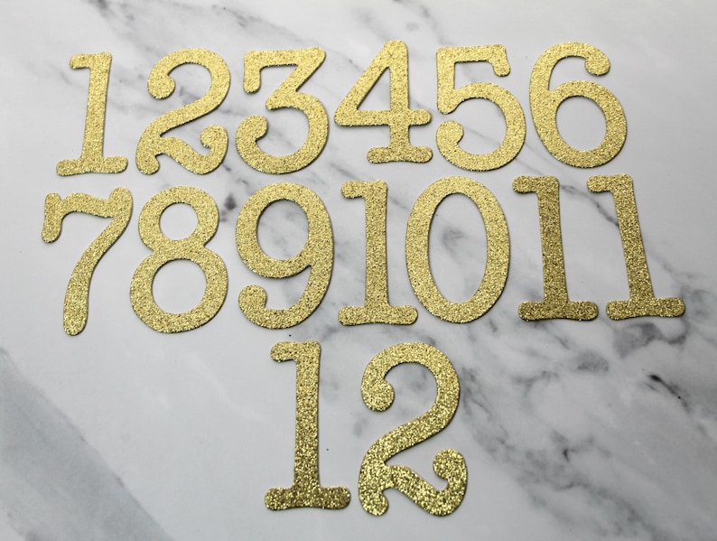 May include: Gold glitter numbers 1 through 12. The numbers are arranged in two rows. The first row has numbers 1 through 6. The second row has numbers 7 through 11. The third row has numbers 1 and 2.